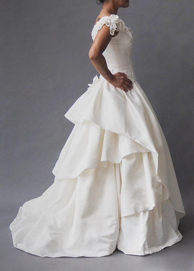 CINDERELLA BRIDAL GOWN Rosette embellished cap sleeves and a bead-encrusted, boned bodice dive into an off-white tiered skirt, inflated by layers of tulle and masses of crinoline. Zip closure masquerading as a row of buttons ensure ease and wearability.