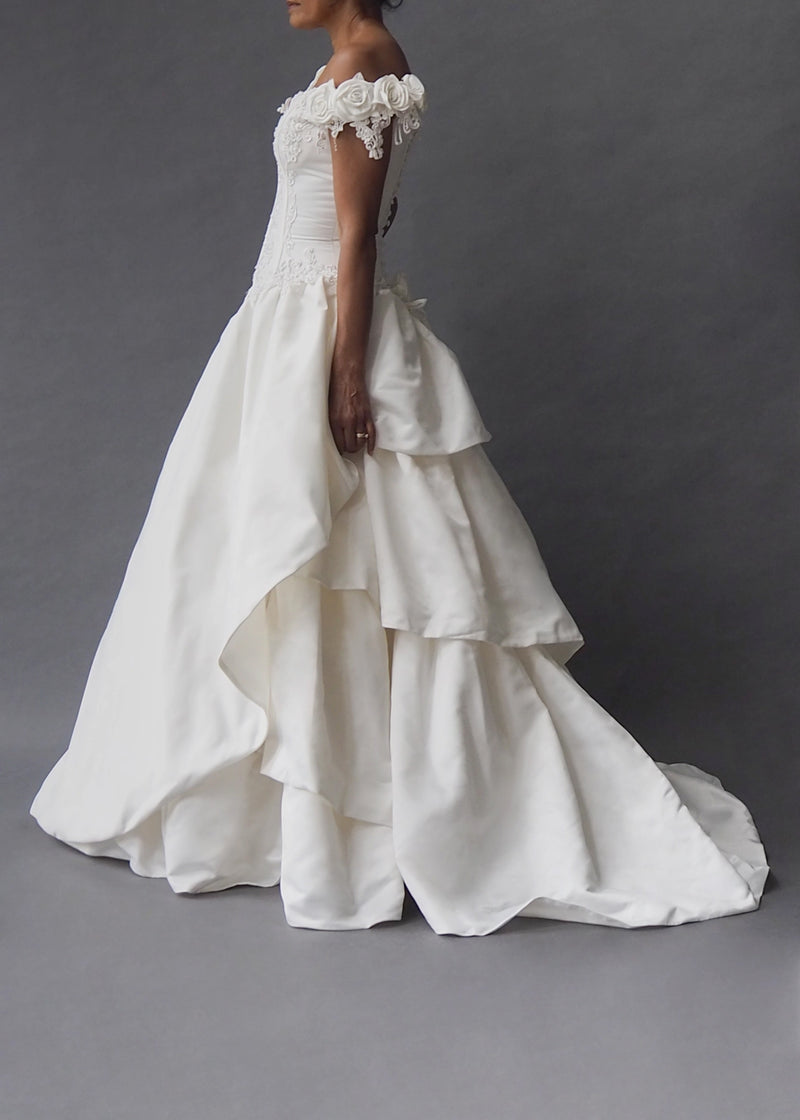 CINDERELLA BRIDAL GOWN Rosette embellished cap sleeves and a bead-encrusted, boned bodice dive into an off-white tiered skirt, inflated by layers of tulle and masses of crinoline. Zip closure masquerading as a row of buttons ensure ease and wearability.